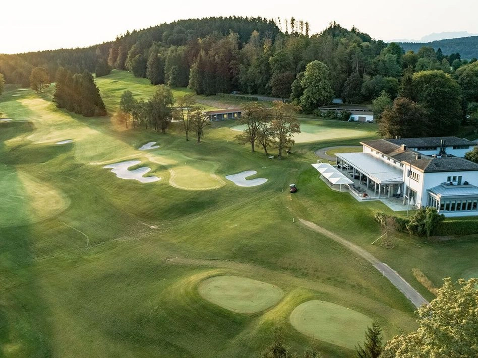 Aerial view of the Golf & Country Club Zürich in Zumikon showing the 18-hole championship course with green fairways and greens, the historic Bauhaus-style clubhouse, and the surrounding hilly landscape above Lake Zurich.